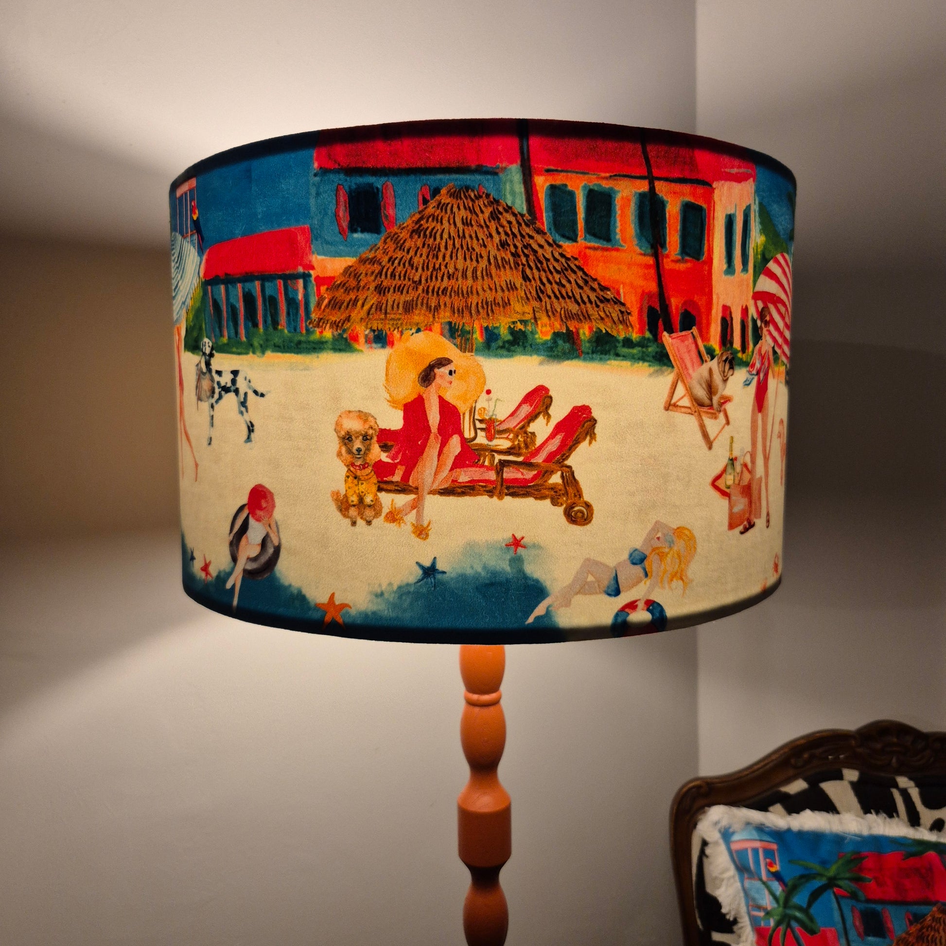 Queen K Riviera Drum Lampshade with colorful beach scene and fringed design on a wooden lamp base
