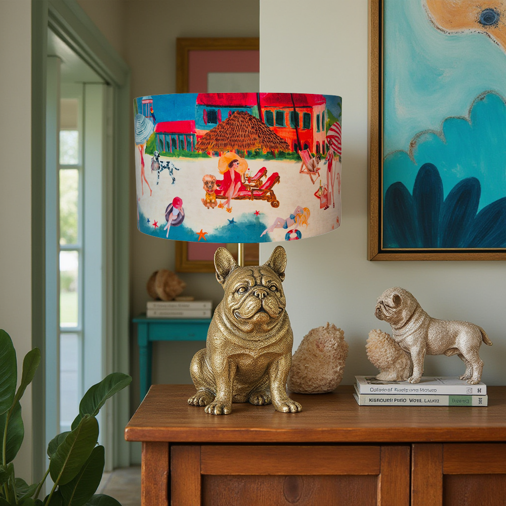 Queen K Riviera Drum Lampshade with colorful beach scene on fringed drum lampshade above gold dog figurine lamp base