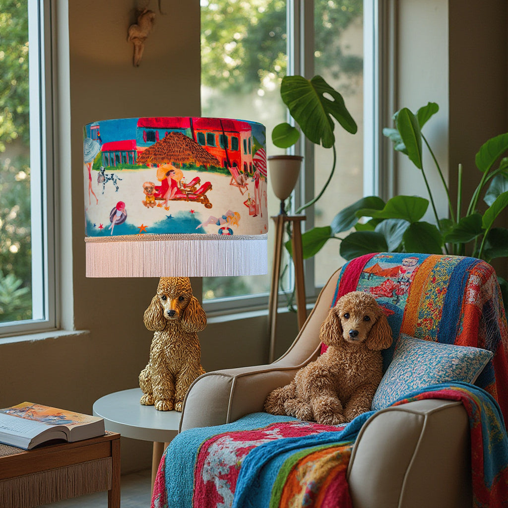 Riviera Fringed Drum Lampshade by Queen K with colorful beach scene and white fringe on gold dog base in cozy living room
